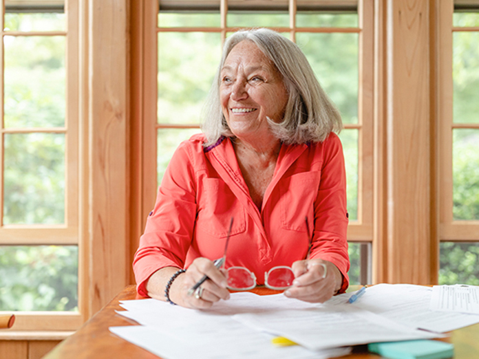 Woman smiling wearing a coral-colored shirt holding a pen and eyeglasses.