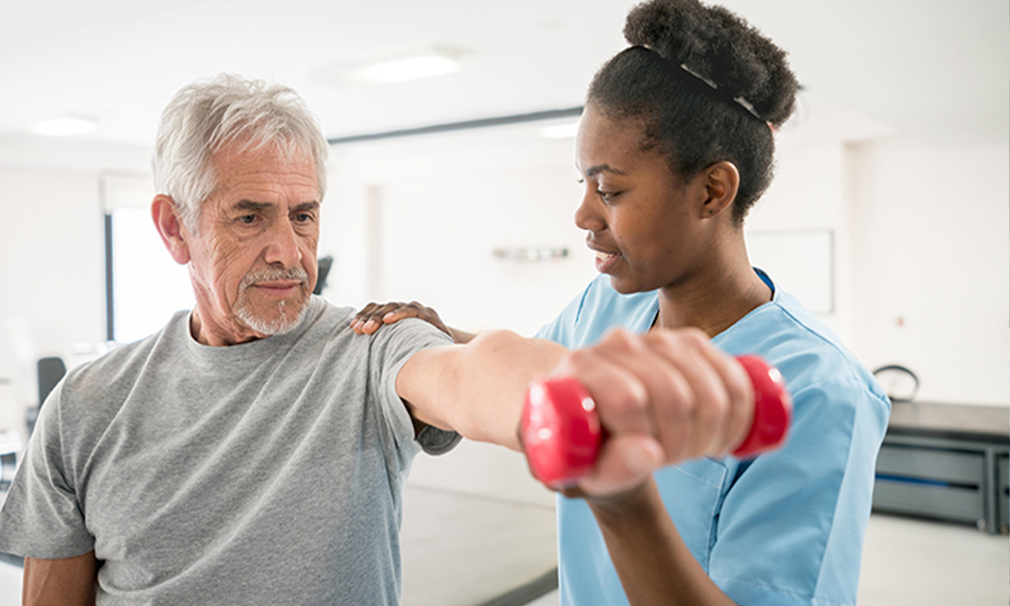 Male wearing a grey t-shirt lifting a weight while a female nurse in blue scrubs helps him.