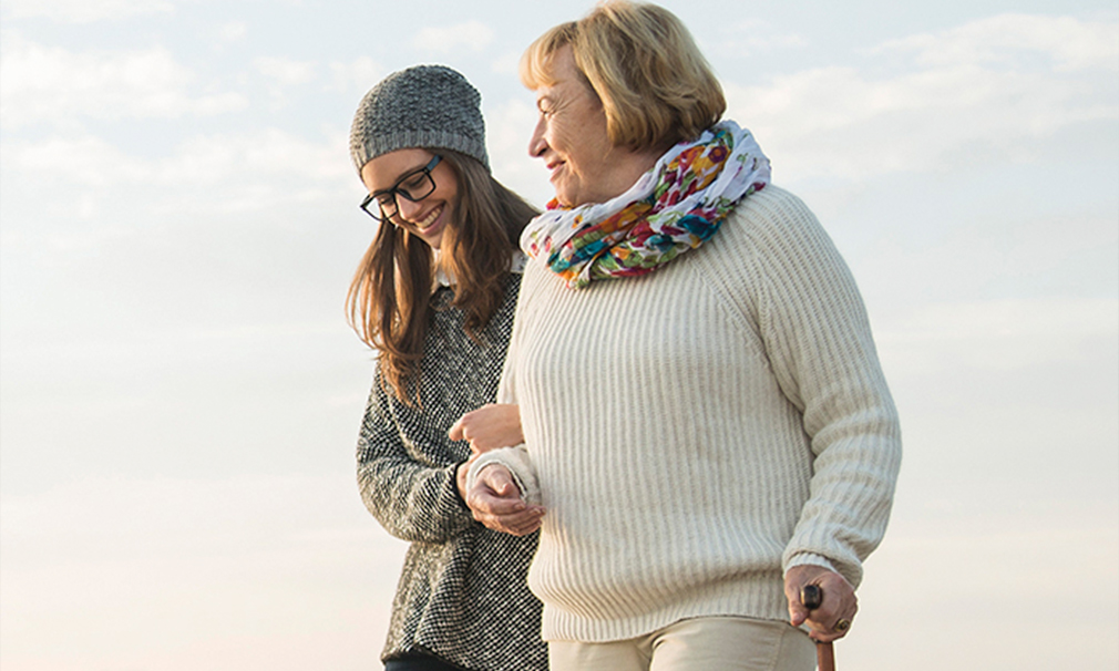 Female wearing a hat and eyeglasses holding a woman's arm as she uses the other arm to walk with a cane.